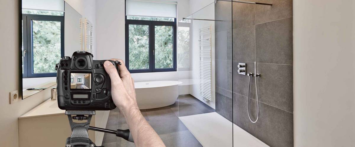 Hands holding a professional camera on tripod taking picture in tiled bathroom with windows towards garden