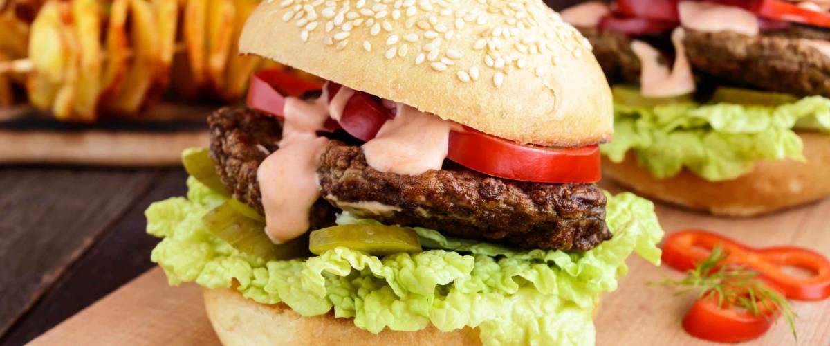 Burgers with liver cutlet, tomatoes, pickles, lettuce, spicy sauce and a soft bun with sesame seeds on a cutting board and potato slices on skewers on a dark wooden background. Close up