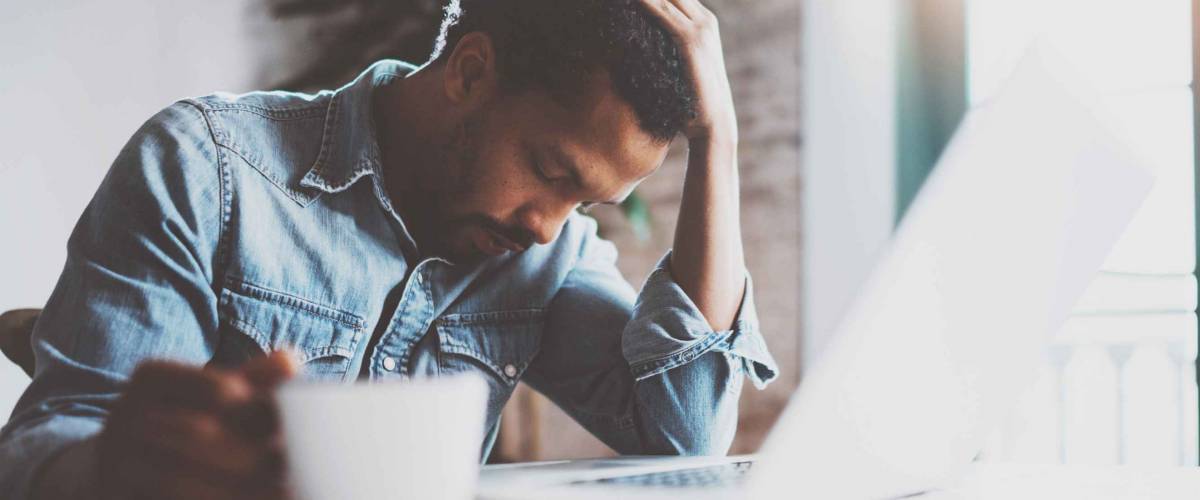 Tired young African man using laptop while sitting at the table on a sunny morning.Concept of people working hard home.Blurred background,flare effect