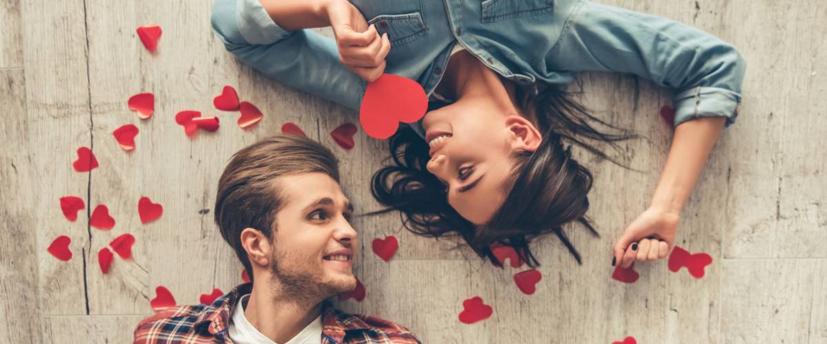 Top view of happy young couple looking at each other and smiling while lying on wooden floor. Girl is holding a red paper heart