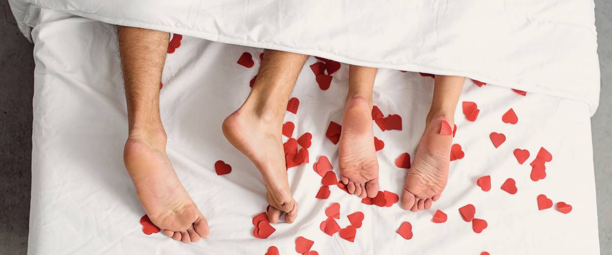 Close up of male and female feet. Loving couple is lying on bed under blanket covered by small red paper hearts