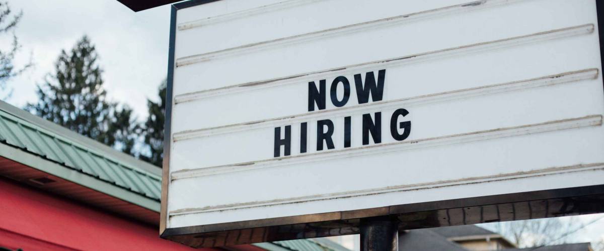 Typical american now hiring sign in black letters over the white background with local small business building