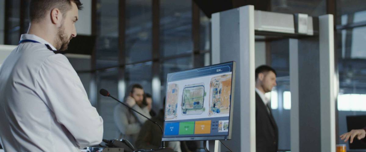 Man in uniform standing at counter at checking point and watching at monitor with x-ray of luggage.