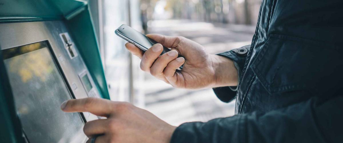 Closeup of male hands using smart phone while typing on ATM, bank machine
