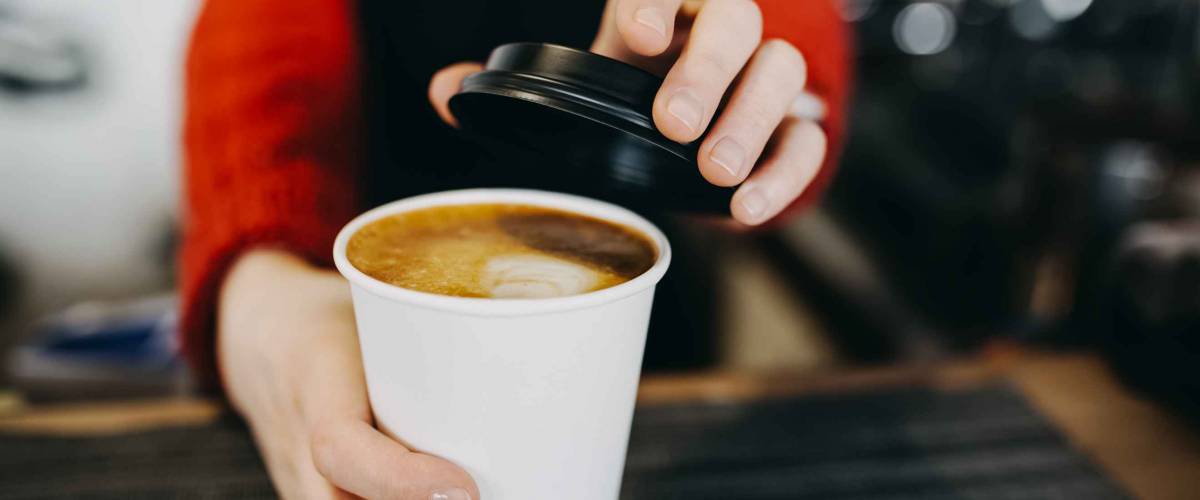 Barista in apron is holding in hands hot cappuccino in white takeaway paper cup. Coffee take away at cafe shop