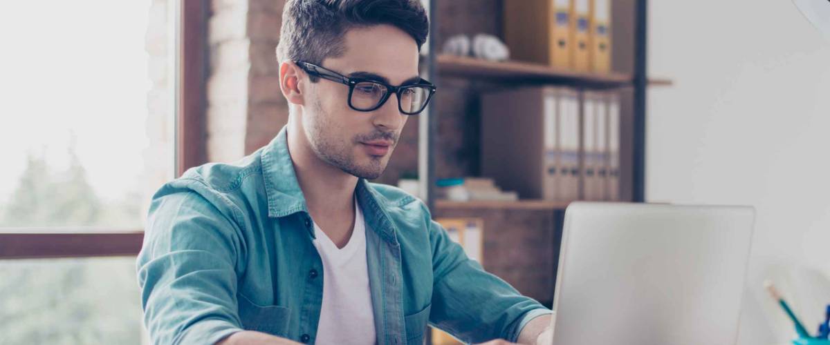 Young handsome minded man in jeans shirt and spectacles sitting at the table and typink a letter to his acquaintance