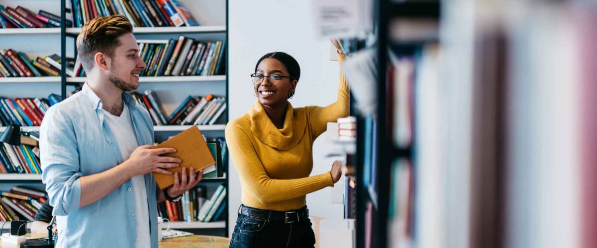 Positive african american female librarian helping caucasian student to search literature book in bookshelf.