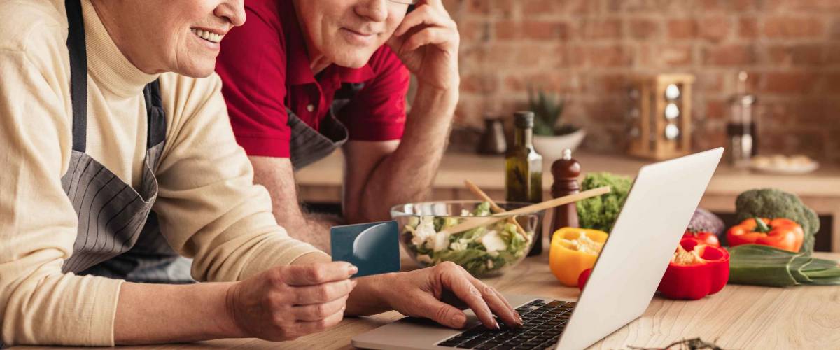 Ordering Food Online. Smiling elderly couple with laptop and credit card purchasing grocery delivery from internet while cooking together in kitchen