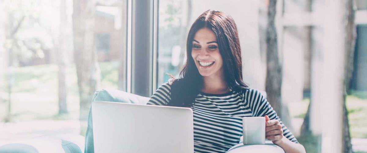 Enjoying time at home. Beautiful young smiling woman working on laptop and drinking coffee while sitting in a big comfortable chair at home
