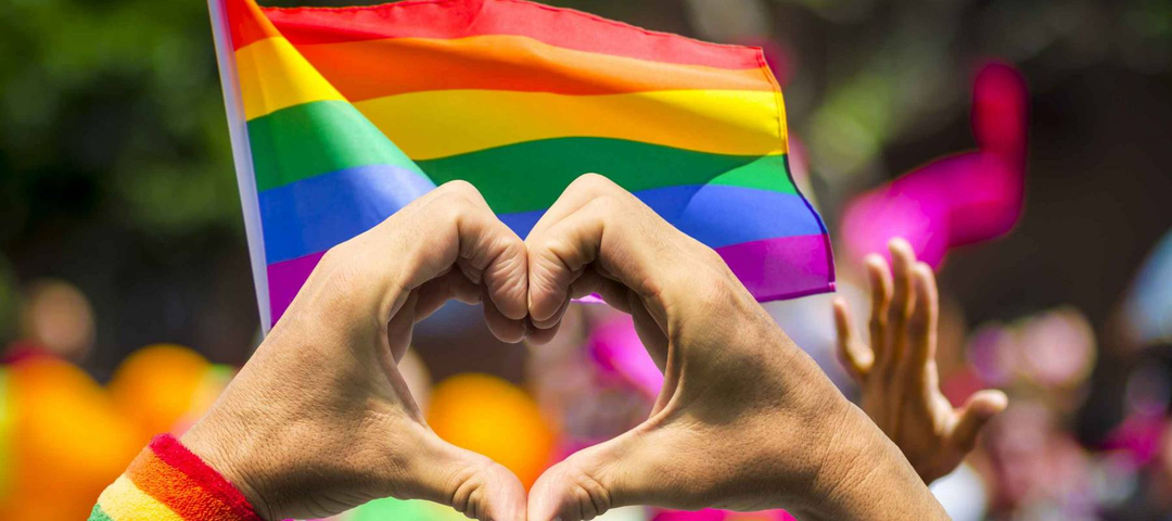 Supporting hands make heart sign and wave in front of a rainbow flag flying on the sidelines of a summer gay pride parade