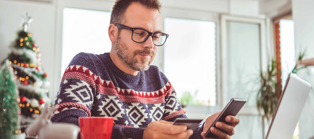 Men wearing blue sweater and eyeglasses holding credit card and using laptop at home office during christmas holidays