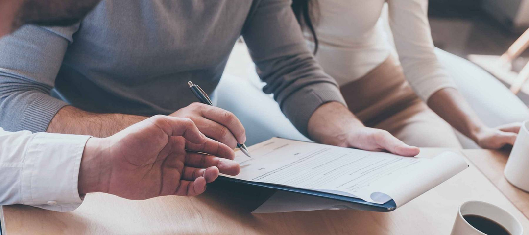 Signing documents. Close-up of confident young man signing some document while sitting together with his wife and another man pointing document