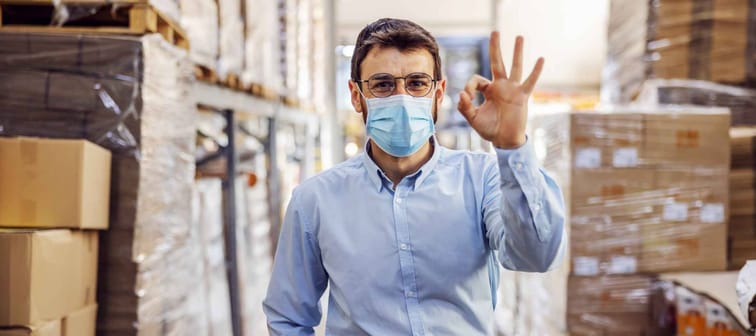 Young businessman with sterile protective mask on standing in warehouse and showing okay sign. Protection from covid-19.