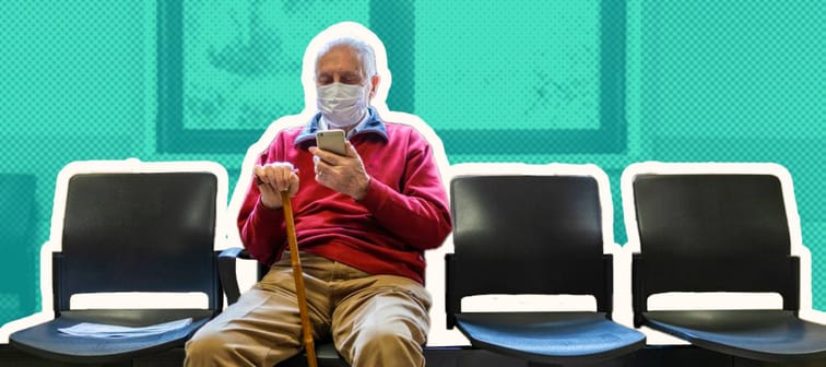 Older man sits wearing a mask in a doctor's office waiting room, looking at his phone.