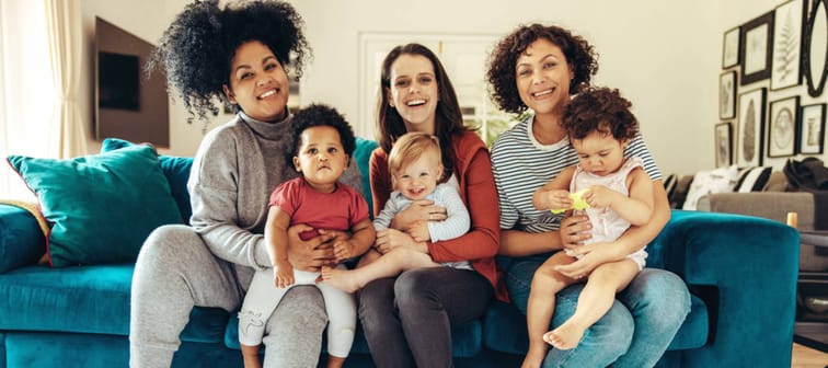 Three moms sit on a couch, smiling and holding babies.