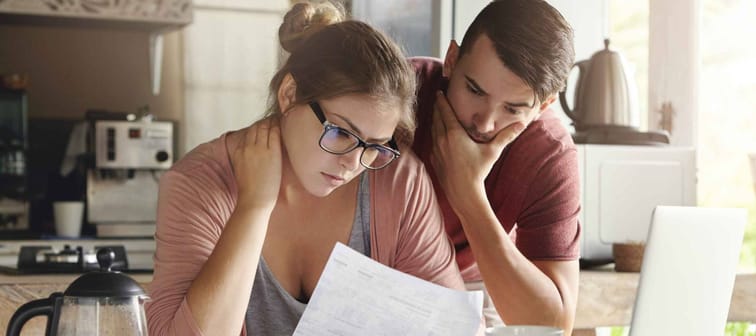 Young couple wondering if they should sell their home when examining all the costs looking at paperwork with calculator, laptop, and coffee