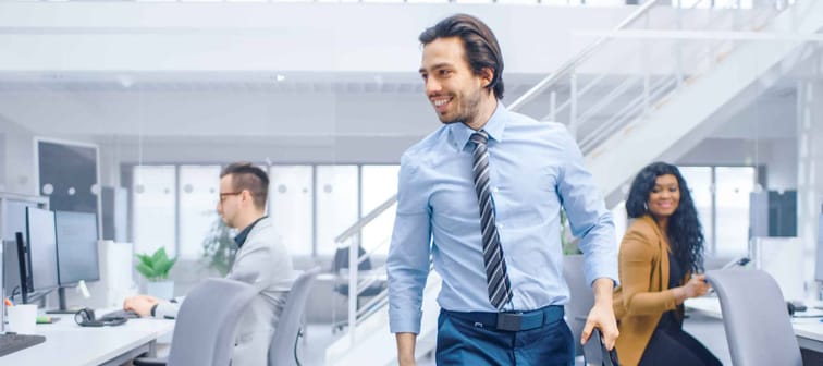 Young Happy Businessman Returns to Work, Walks Into Bright Modern Office, Greets Everybody, Takes Place at His Desktop Computer.