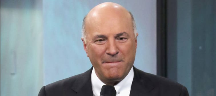TV personality Kevin O'Leary on the set of a television show, sitting on a stool and holding a microphone, pursing his lips.
