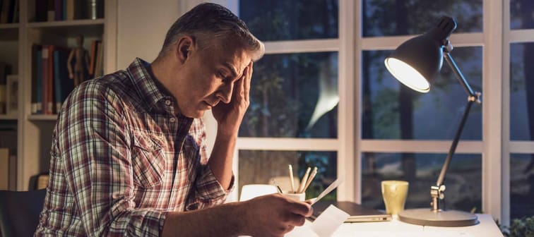 Desperate mature man checking his domestic bills at home, he is holding an invoice and touching his head