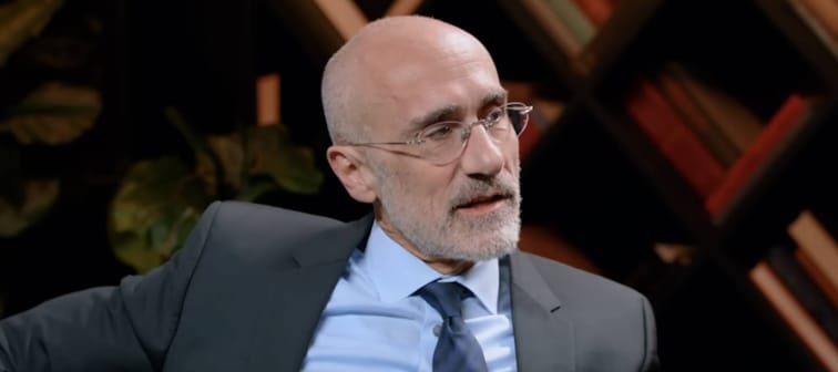 Man in glasses and suit sits looking to the side of the camera, speaking expressively.