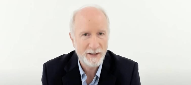 Older man in a suit, looking directly at the camera and talking.