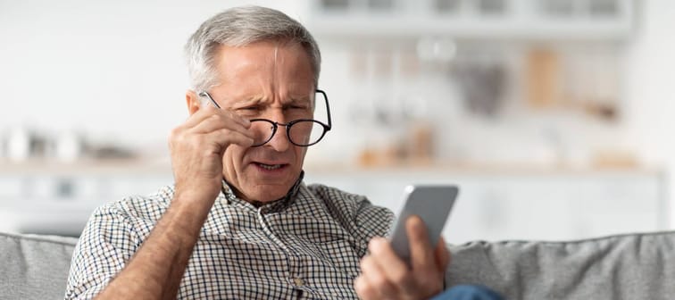 Senior Man Squinting Eyes Reading Message On Phone Wearing Eyeglasses, confused Sitting On Couch At Home.