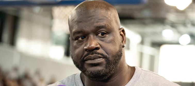 Shaquille O'Neal speaking into a microphone, looking off to the side and sad.