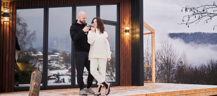 Happy woman and man clinking glasses of wine and celebrating together on winter day in mountains. Couple in love toasting enjoying alcoholic drink outside wooden house with panoramic window.
