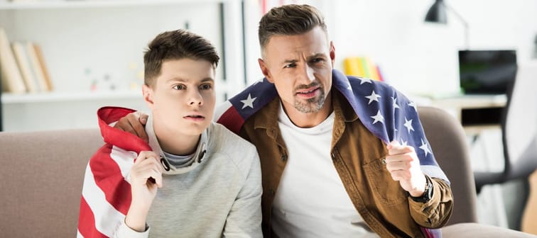 serious father and teen son wrapped in united states flag sitting on sofa and watching sport at home
