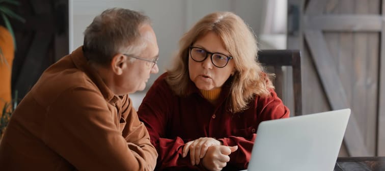Two older people sitting at a table with laptop, looking serious.