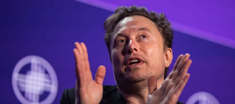 Elon Musk seen on stage, blue background behind him, hands held upwards and looking up.