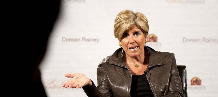 Suze Orman speaks leaning to one side, sitting in a chair, with her arm held up, speaking dramatically.