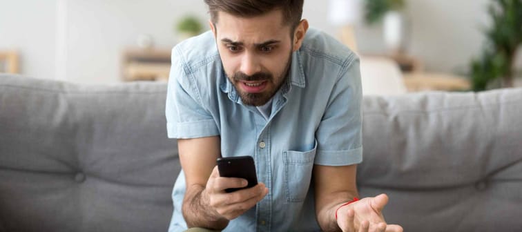 Confused millennial man sitting alone on couch in living room at home looking at smartphone