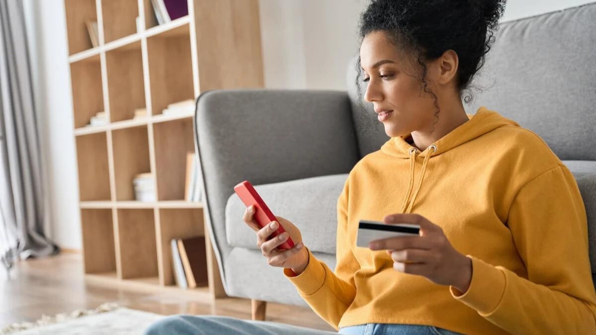 Young adult African American female consumer holding credit card and smartphone sitting on floor at home doing online banking transaction. E commerce virtual shopping, secure mobile banking concept.