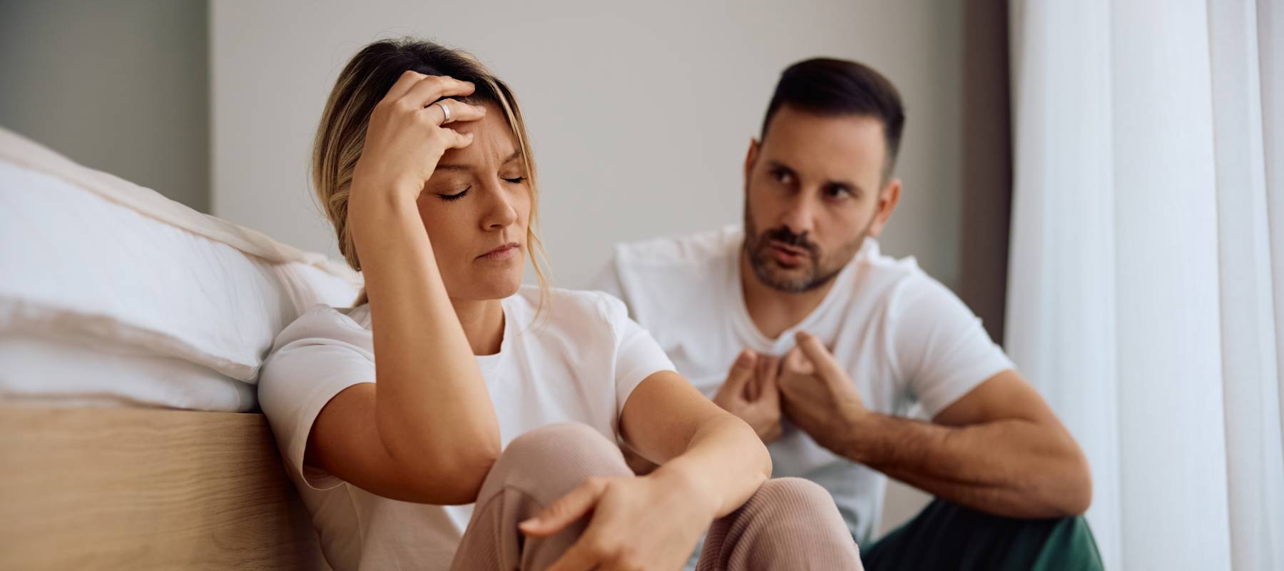 Mid-30s woman sitting by bed, holding her head in frustration as her male partner tries to explain himself.