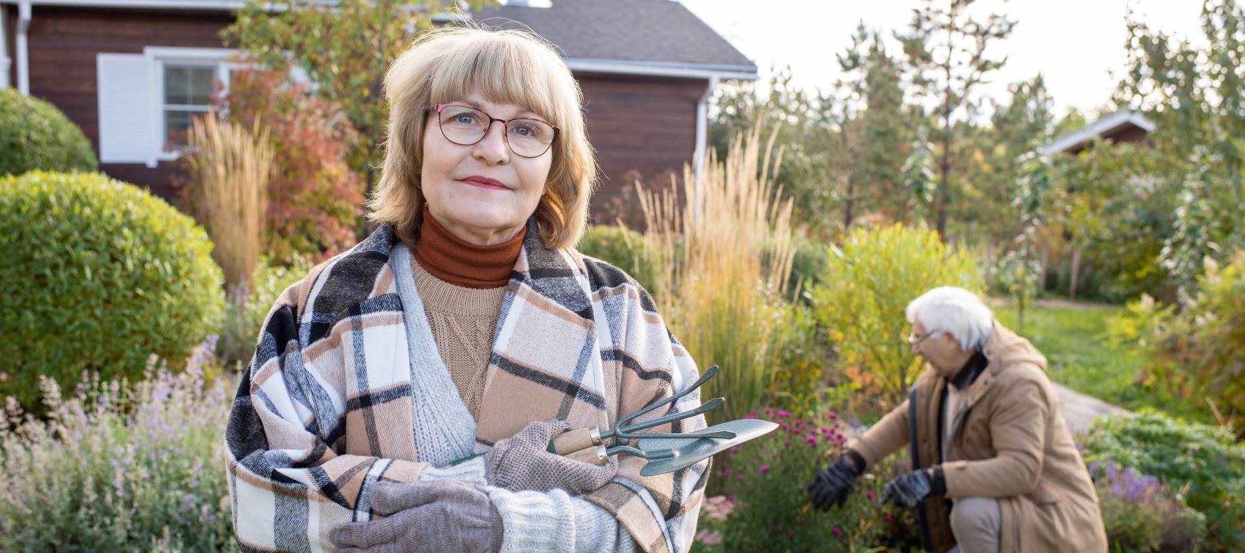 Older woman looking at camera holding gardening tools with man in the background