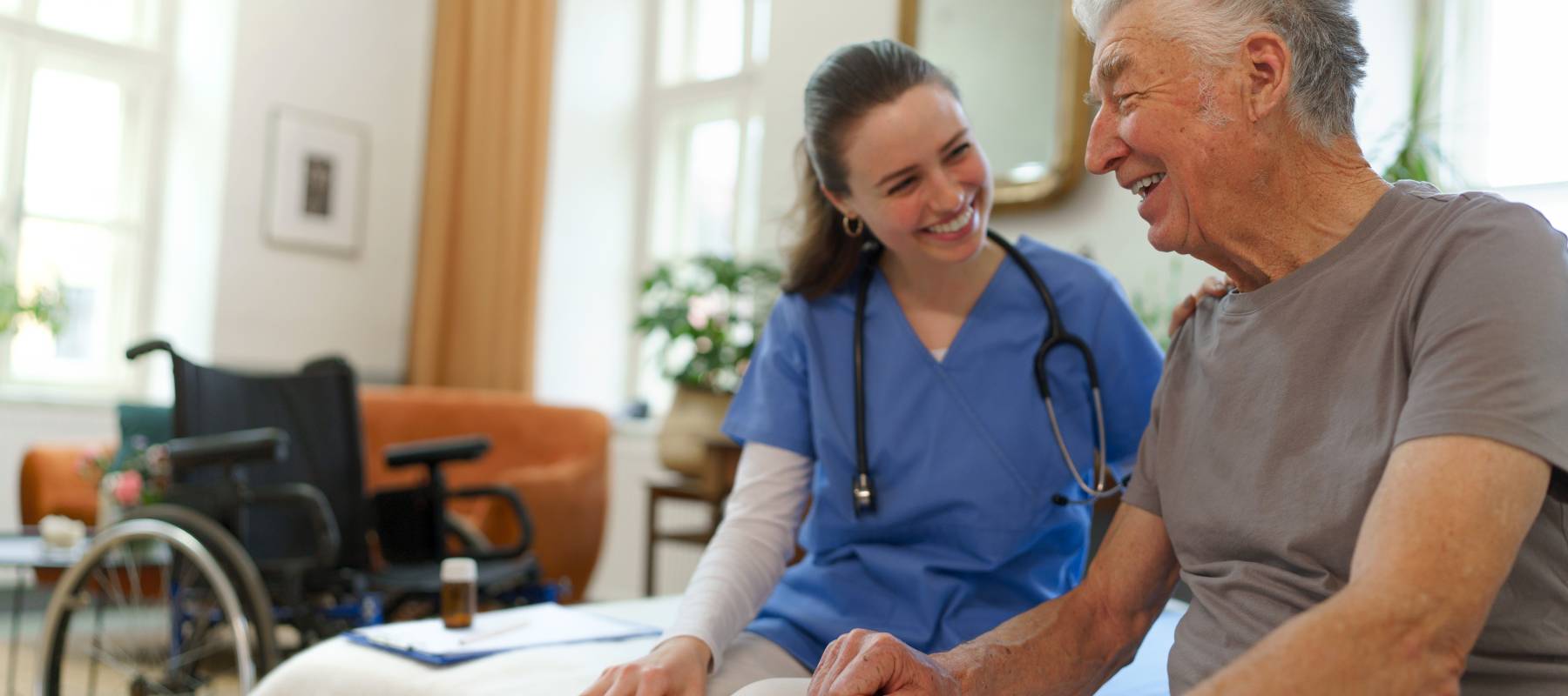 A senior at a nursing home sitting on the bed with a nurse.