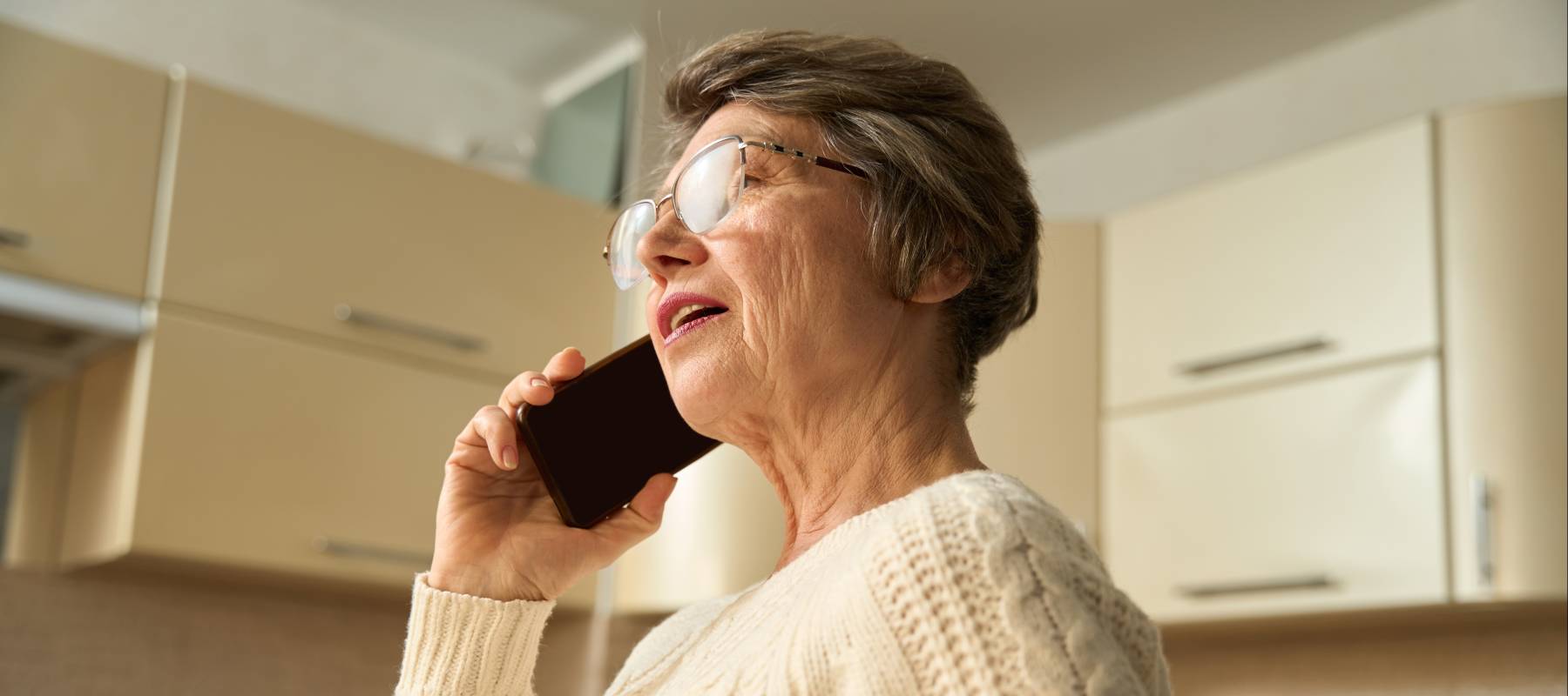 Old woman with glasses standing in the kitchen, holding smartphone and having call