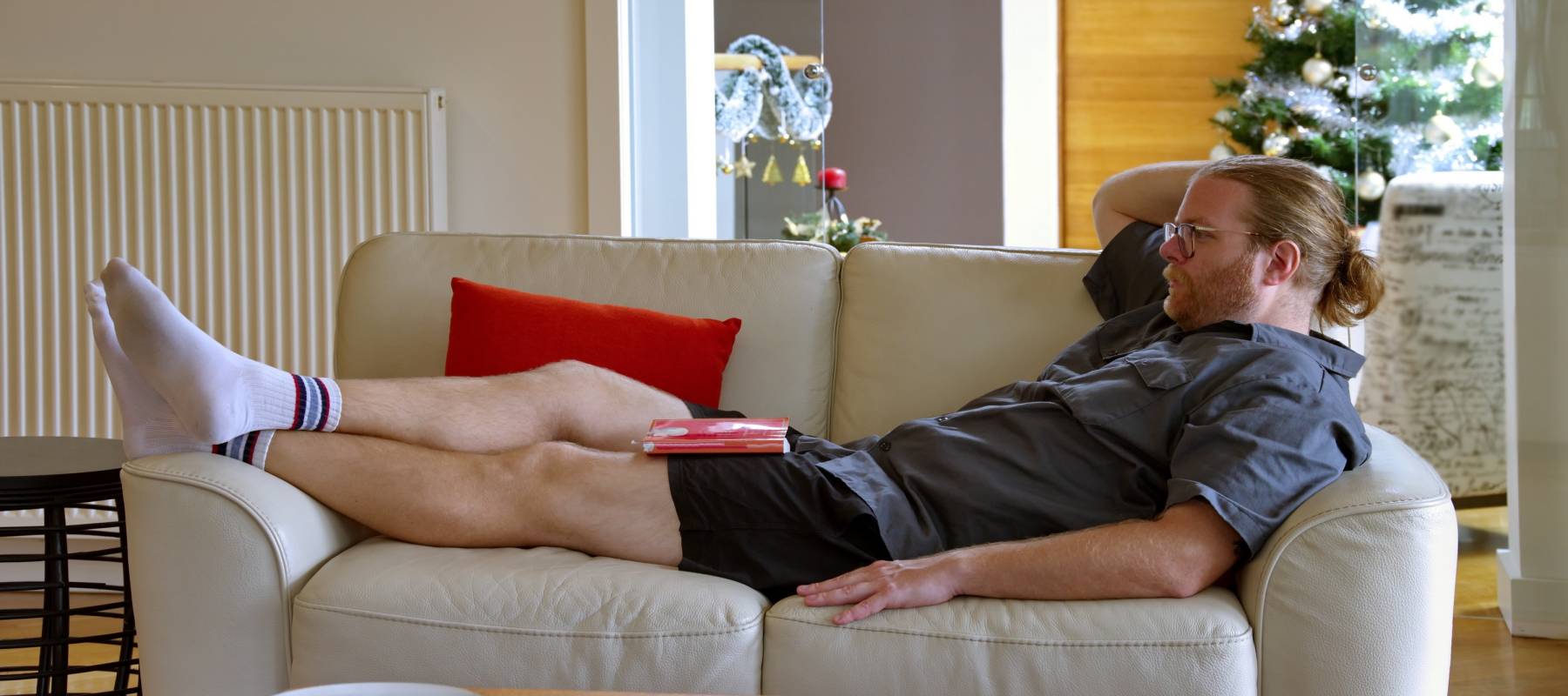 Bespectacled man with a ponytail, laying on the couch with a plate next to him and a book on his lap, staring off into the distance.