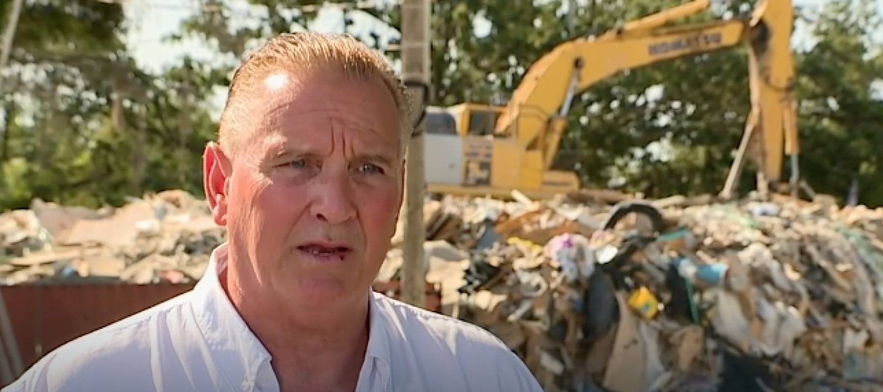 Man speaks to camera in front of a mound of debris.