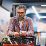 Bespectacled man with gray hair and beard leaning on his shopping cart and holding a receipt with a distant look in his eyes.