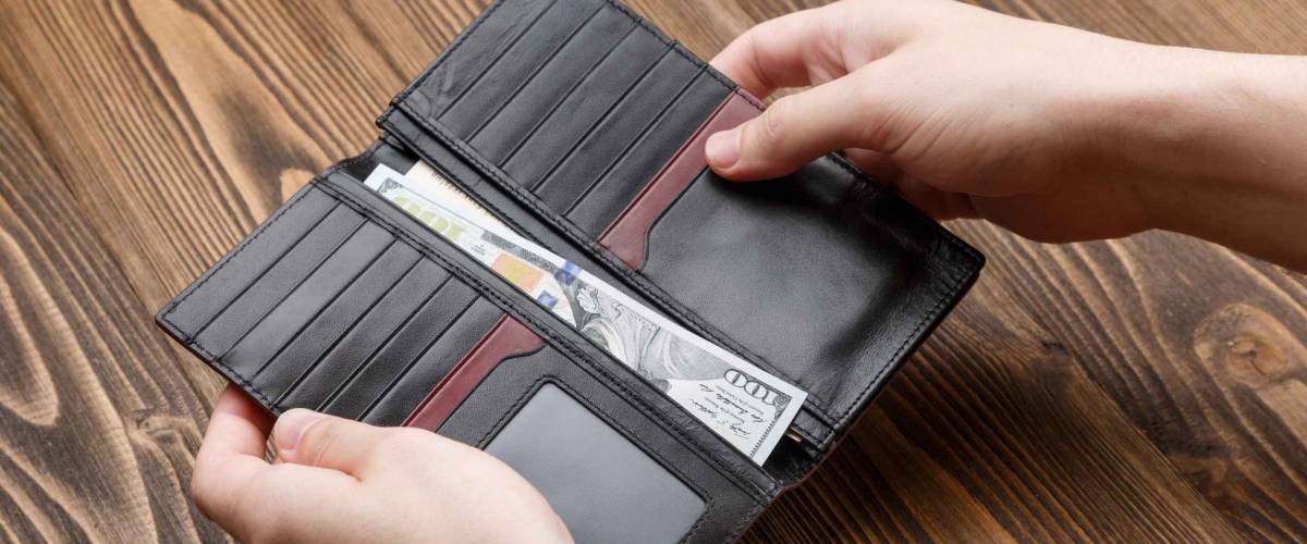 man is putting 100 dollar banknote in a black leather wallet over dark wooden background. Close up shot of hands and wallet