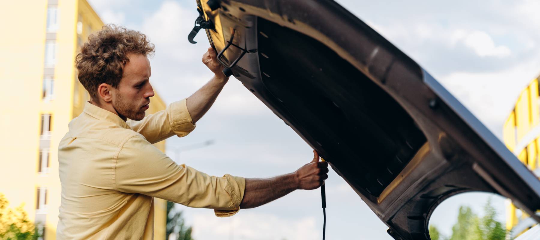 Frustrated man propping open car hood in city landscape.