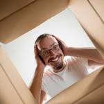 The surprised man unpacking, opening carton box and looking inside. The package, delivery, surprise, gift lifestyle concept. Human emotions and facial expressions concepts