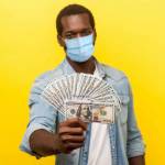 Portrait of joyous wealthy man with surgical medical mask holding out dollar bills at camera, boasting money won in lottery. indoor studio shot isolated on yellow background