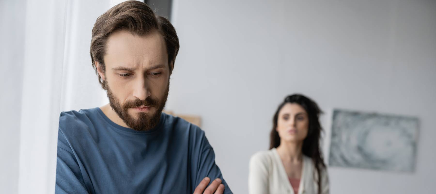 Contemplative man standing cross-armed by a window with his female partner in the background, looking at him from behind.