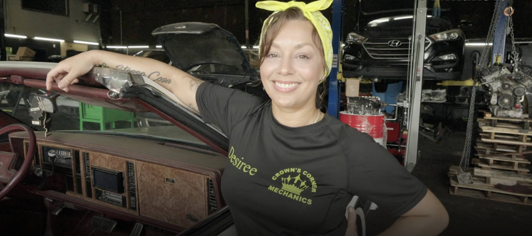 Georgia mom proudly standing and smiling in her auto mechanic shop, in a black company t-shirt and a lemon yellow bandanna tied in a bow on top of her head.