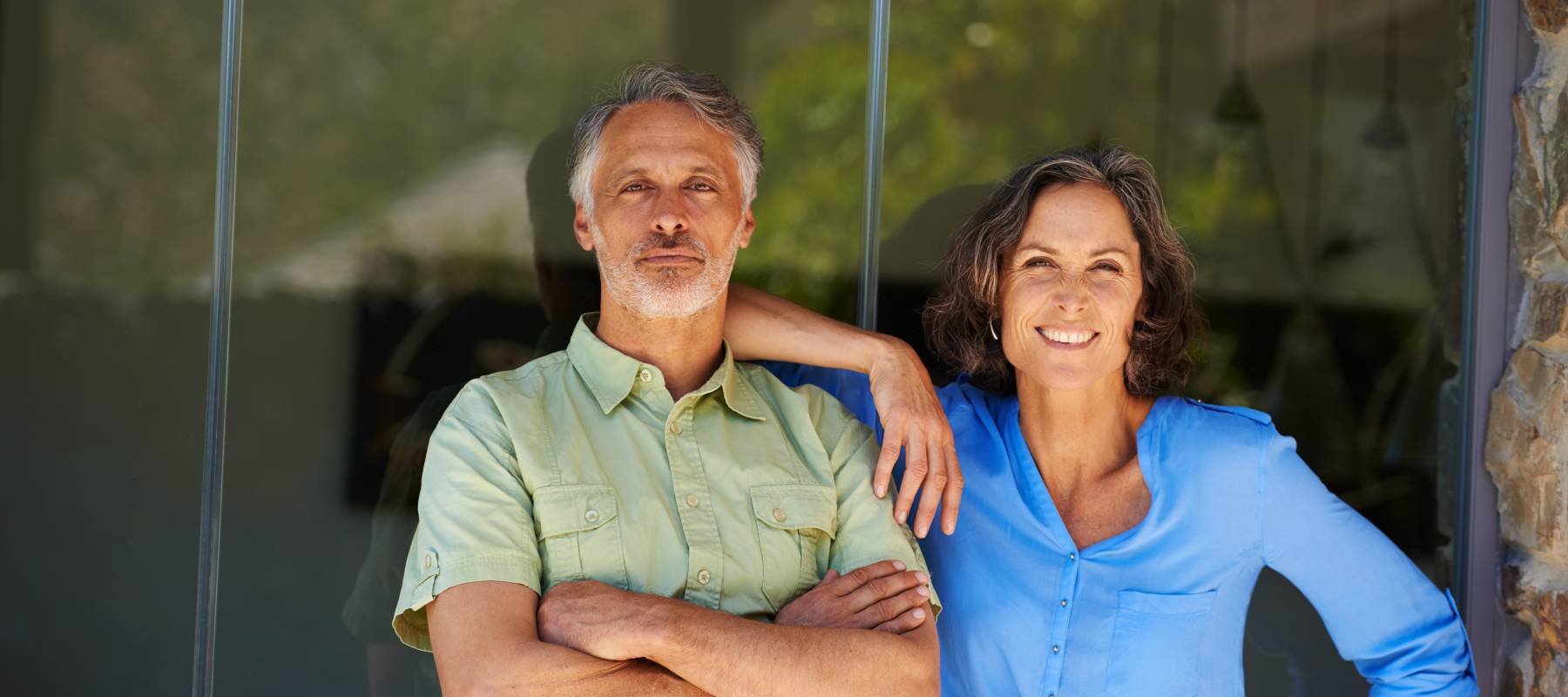 Older-aged couple standing by a sliding glass door with confident wide smiles.