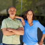 Older-aged couple standing by a sliding glass door with confident wide smiles.