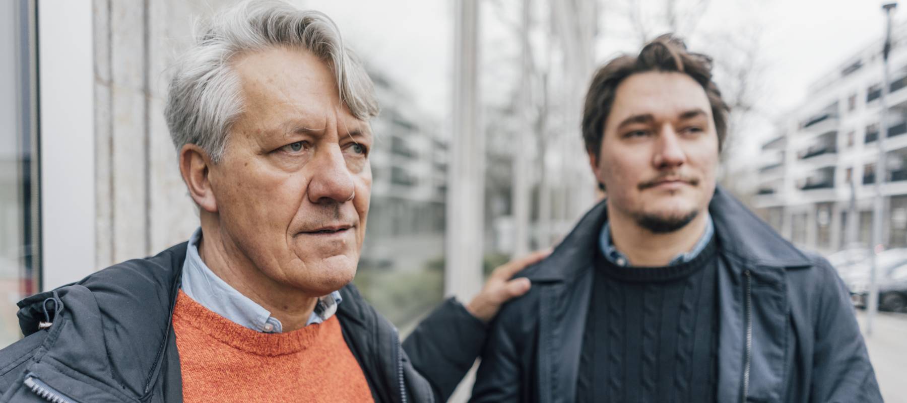 Older gentleman in focus and looking off into the distance, with a younger man in the background and out of focus.